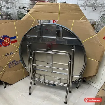 Bàn tròn inox Hwata BT10 gấp chân dựng đứng, phía sau là thùng hàng ghi quy cách 1000 mm