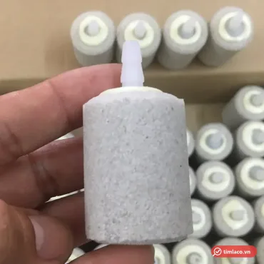 Cục sủi oxy mịn hình trụ màu xám trắng có đầu nối nhựa, phía sau là nhiều viên cùng loại xếp trong thùng