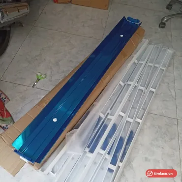 Máng đèn đôi chóa inox 1m2 đặt trên thùng carton, chóa còn màng bảo vệ màu xanh (ảnh minh hoạ)