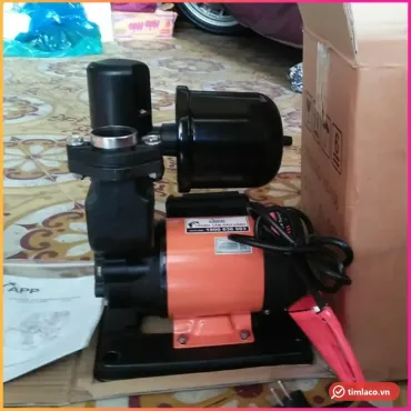 Máy bơm tăng áp APP động cơ vỏ cam, đầu bơm màu đen, bình tích áp phía trên, đặt cạnh thùng hàng (ảnh minh hoạ dòng hàng)
